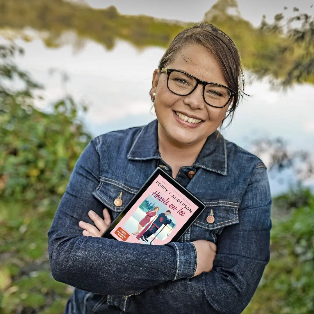Poppy J. Anderson, sie lächelt sympathisch in die Kamera, im Hintergrund sind unscharfe Pflanzen und ein See zu erkennen