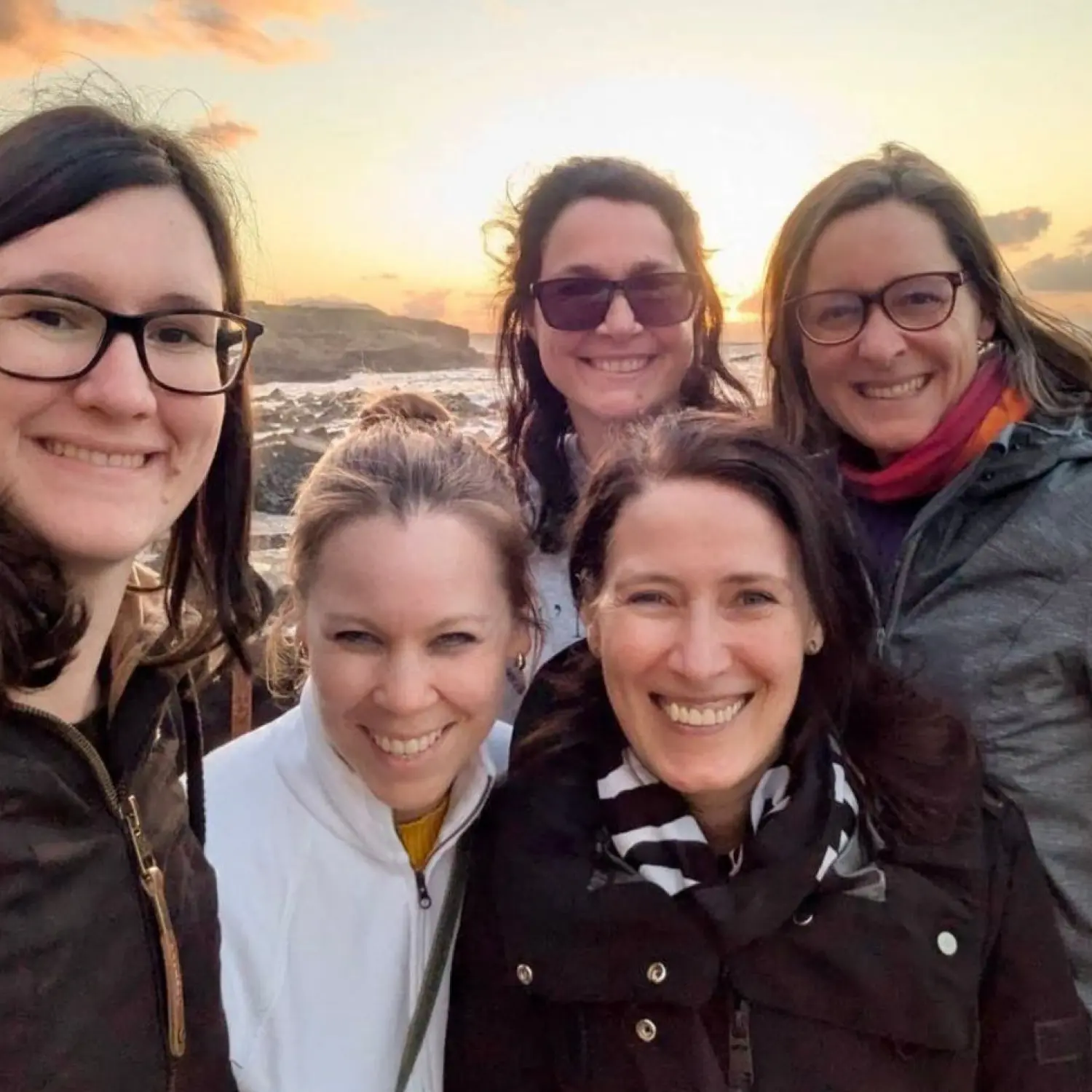 Eine Gruppe von 5 Frauen, die gemeinsam ein Bild bei Sonnenuntergang am Strand machen
